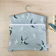 peg bag with zip closure a third of the way down. bag hangs using a coat hanger inserted inside with just the hook showing at the top. swallows are detailed and are flying on a blue background
